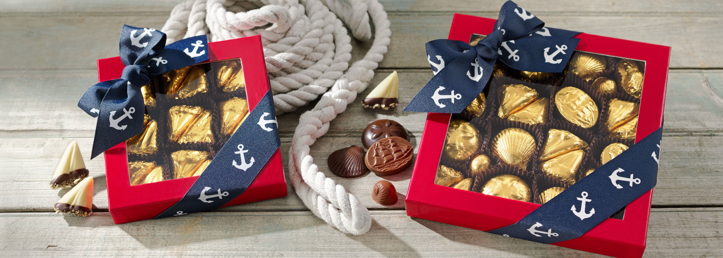 Two gift boxes with chocolate and a decorative anchor ribbon on a wooden surface. Boxes feature gold foiled nautical collection.